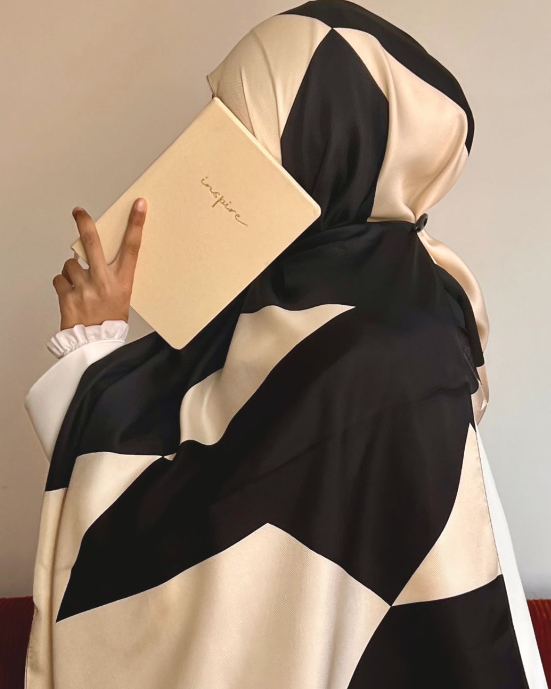 Femme musulmane en hijab à motifs géométriques noirs et écru en soie 100%, vêtue d’une abaya blanche avec un livre à la main dans un studio parisien. 