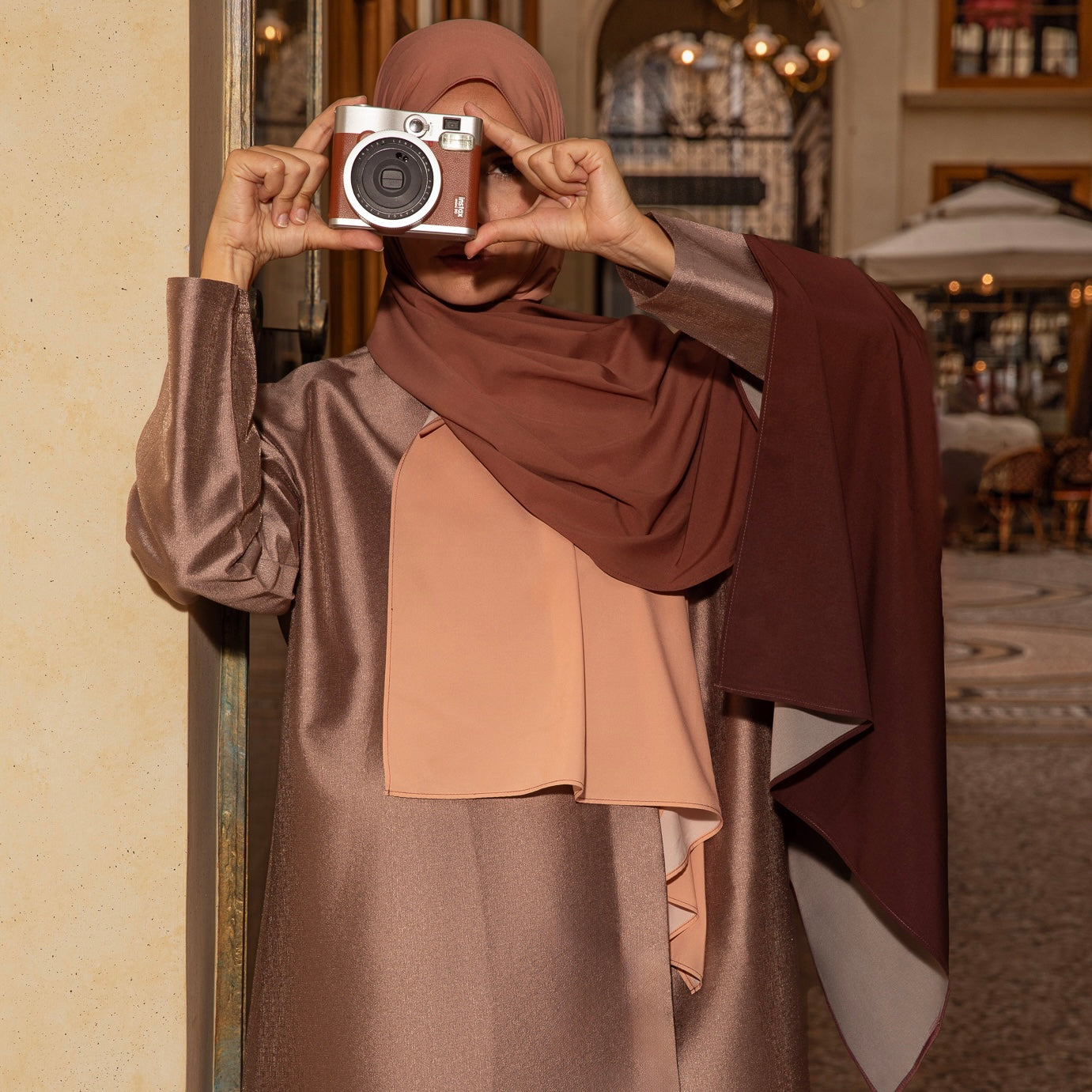 Femme voilée élégante portant un hijab dégradé Cannelle Corsée, dans un décor raffiné, tenant un appareil photo – Collection Mimi Kiseki.
