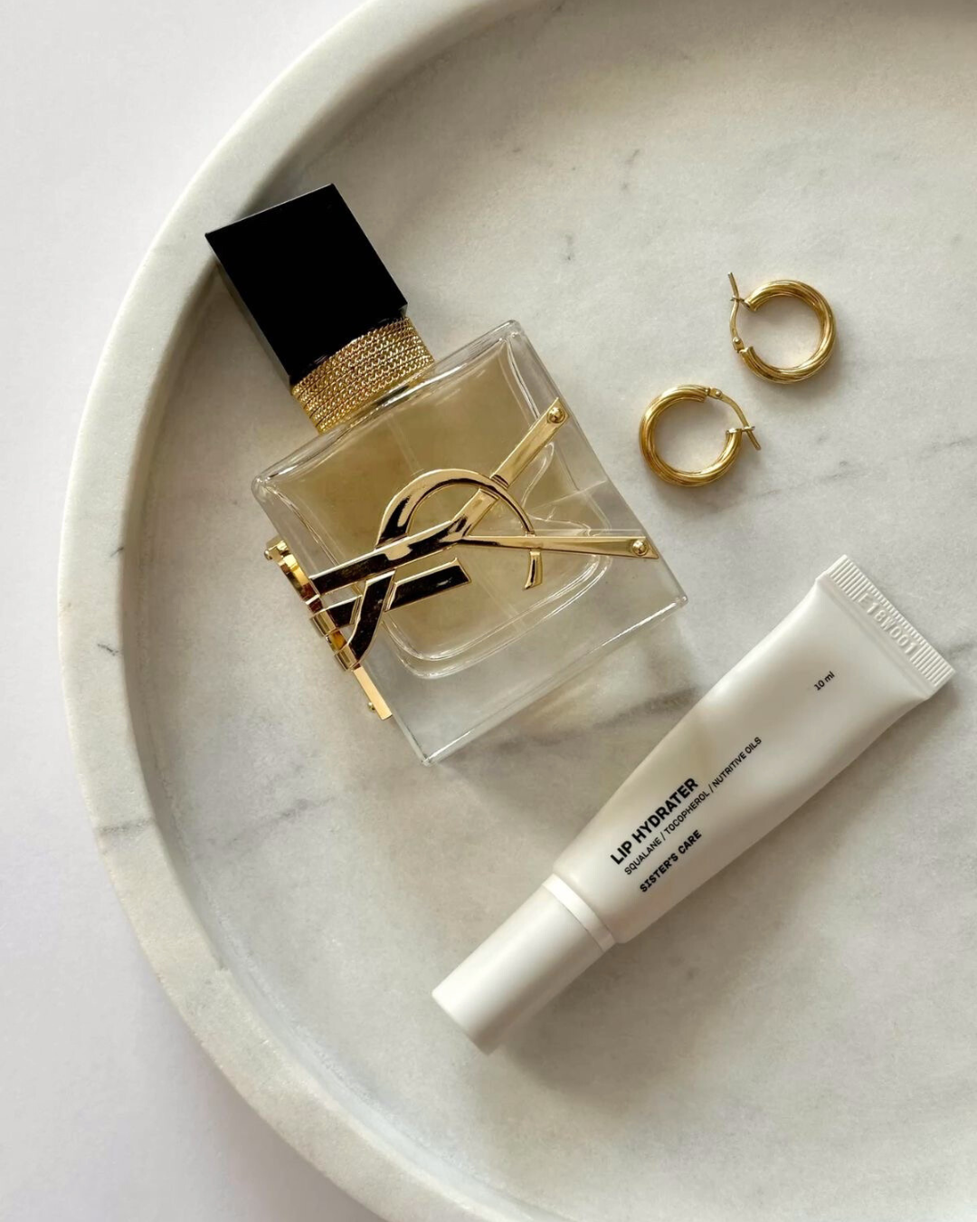 Collaboration Mimi Kiseki x Marinoush.K – mise en scène élégante avec parfum Yves Saint Laurent, boucles d’oreilles dorées et baume hydratant sur plateau en marbre blanc, illustrant l’univers mode modeste haut de gamme et l’élégance holistique. »