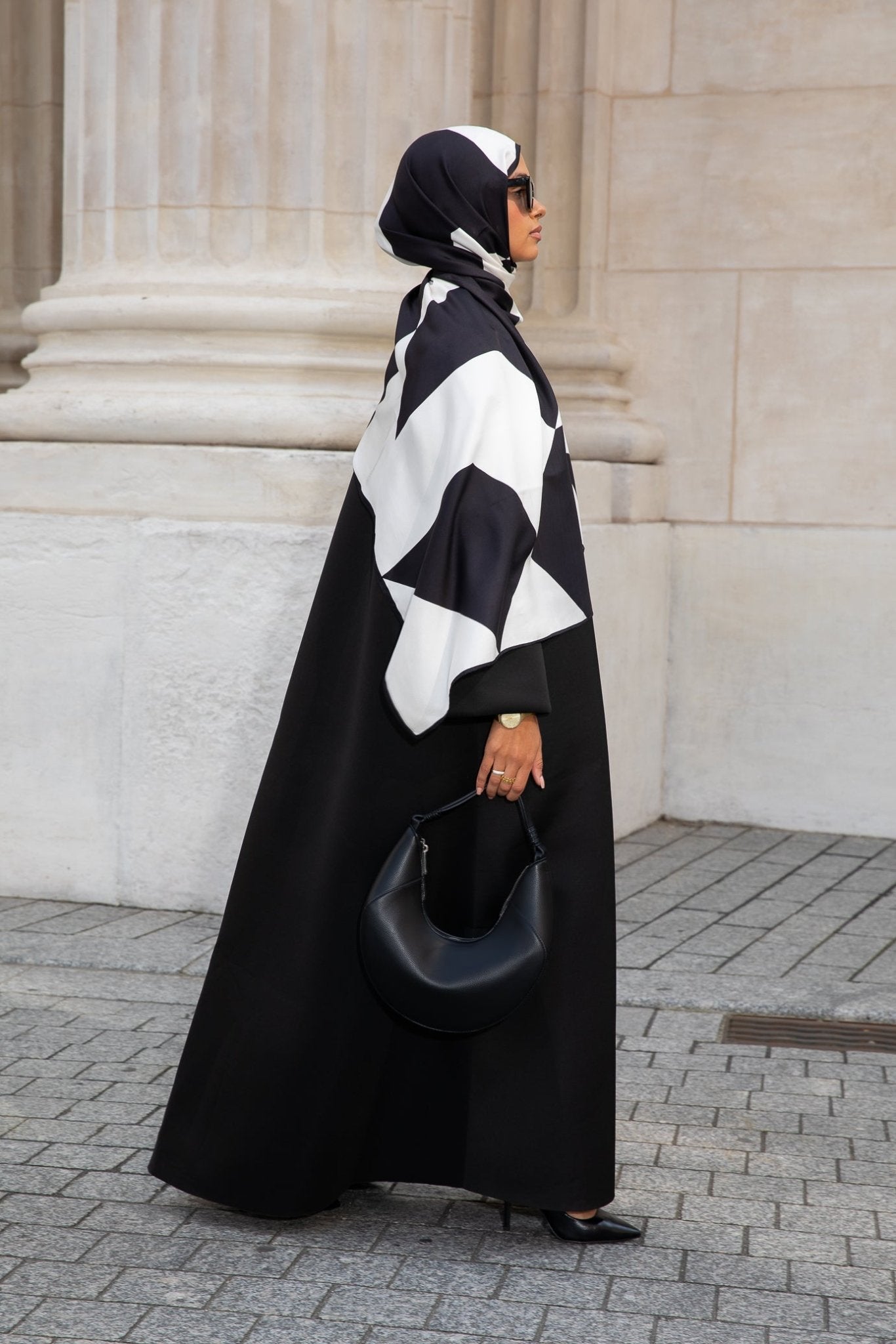 Femme en hijab géométrique noir et blanc Audacy Mimi Kiseki, en tenue noire, posant devant une architecture élégante, parisienne avec un sac structuré noir pour un look affirmé et minimaliste.