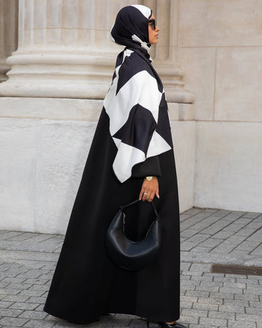 Femme en hijab géométrique noir et blanc Audacy Mimi Kiseki, en tenue noire, posant devant une architecture élégante, parisienne avec un sac structuré noir pour un look affirmé et minimaliste.