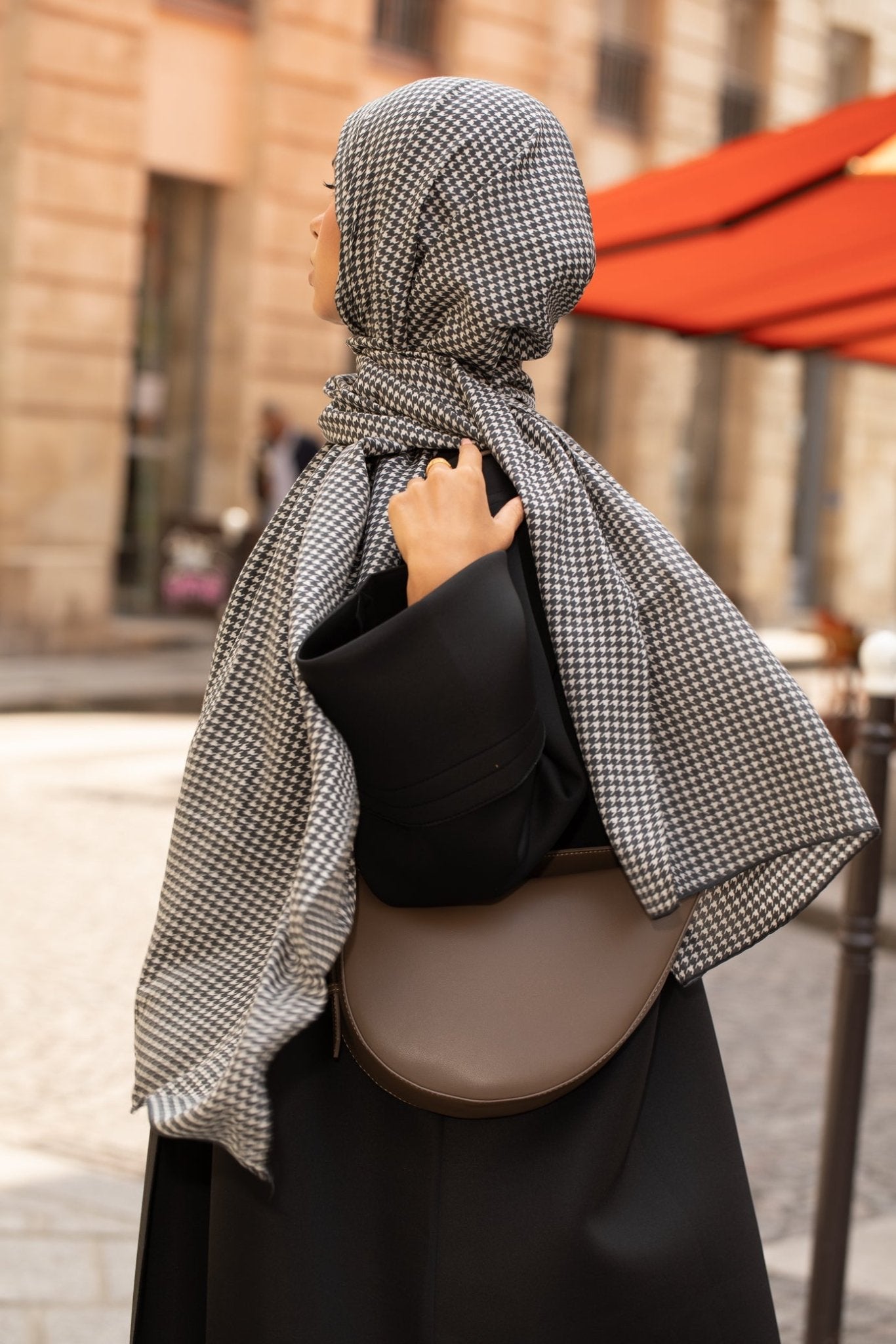 Femme en hijab pied-de-poule Infinity Mimi Kiseki, photographiée de profil en extérieur, portant une abaya noire élégante et un sac arrondi beige, style raffiné et urbain.