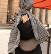Femme en hijab pied-de-poule Infinity Mimi Kiseki, photographiée de profil en extérieur, portant une abaya noire élégante et un sac arrondi beige, style raffiné et urbain.