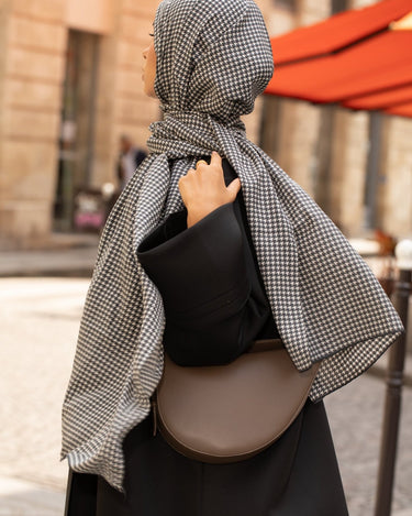 Femme en hijab pied-de-poule Infinity Mimi Kiseki, photographiée de profil en extérieur, portant une abaya noire élégante et un sac arrondi beige, style raffiné et urbain.